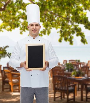 cooking, profession, advertisement and people concept - happy male chef cook showing and holding blank menu board over restaurant lounge on beach