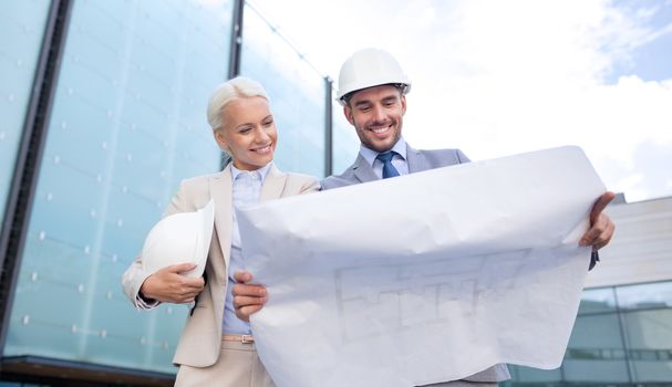 business, partnership, architecture and people concept - smiling businessman and businesswoman with blueprint and helmets on city street