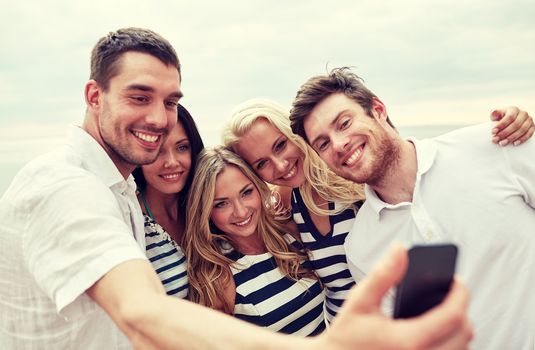 summer, sea, tourism, technology and people concept - group of smiling friends with smartphone on beach photographing and taking selfie