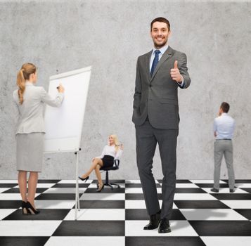 business, people, success, teamwork and strategy concept - smiling young businessman showing thumbs up gesture standing on checkerboard pattern floor over gray background and businesspeople
