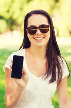 lifestyle, summer vacation, advertisement, technology and people concept - smiling young girl showing black blank smartphone screen and sitting on grass in park