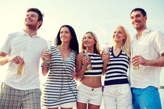 summer, holidays, tourism, drinks and people concept - group of smiling friends with bottles drinking beer or cider on beach
