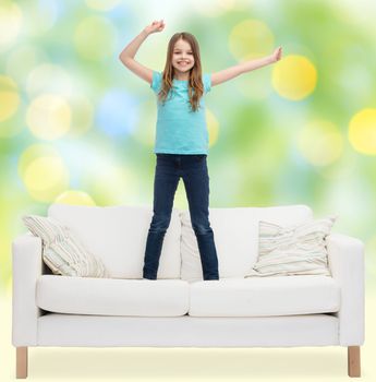 home, leisure, people and happiness concept - smiling little girl jumping and dancing on sofa over green lights background