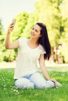 lifestyle, summer, vacation, technology and people concept - smiling young girl with smartphone making selfie and sitting on grass in park