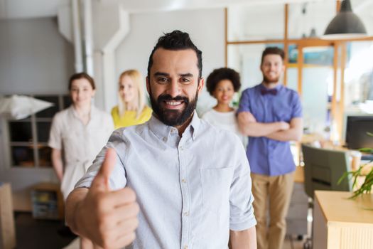 business, startup, people, success and teamwork concept - happy young man with beard over creative team showing thumbs up in office