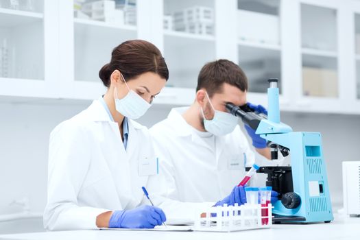 science, chemistry, technology, biology and people concept - young scientists with test tube and microscope making research in clinical laboratory and taking notes