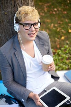 music, technology , drinks and leisure concept - happy teenage boy in headphones with tablet pc computer drinking coffee from paper cup at park