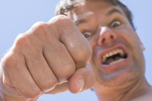 Closeup low angle of vicious angry Caucasian man throwing punch down at camera with sunny blue sky background