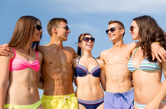 friendship, sea, summer vacation, holidays and people concept - group of smiling friends wearing swimwear and sunglasses talking and laughing on beach