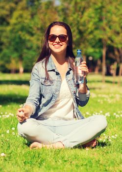 lifestyle, summer, vacation, drinks and people concept - smiling young girl in sunglasses with bottle of water sitting on grass in park