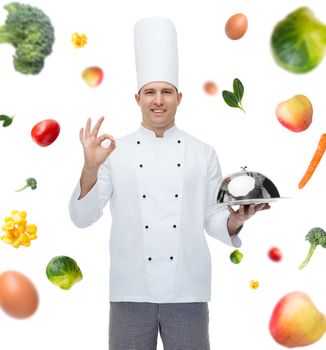 cooking, profession, gesture, vegetarian diet and people concept - happy male chef cook holding cloche and showing ok sign over falling vegetables background