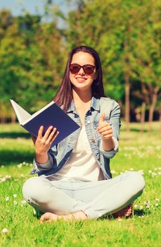 lifestyle, summer vacation, education, gesture and people concept - smiling young girl with book showing thumbs up and sitting in park