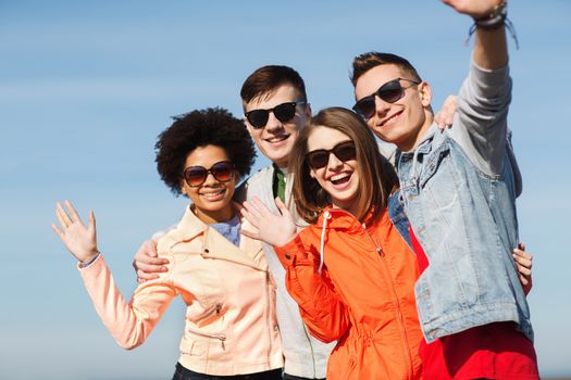 friendship, tourism, travel and people concept - group of happy teenage friends in sunglasses hugging and waving hands outdoors