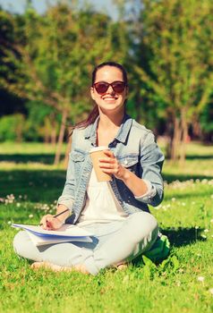 lifestyle, summer vacation, education, drinks and people concept - smiling young girl writing to notebook and drinking coffee in park