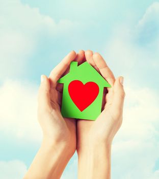 charity, real estate and family home concept - closeup picture of female hands holding green paper house with red heart