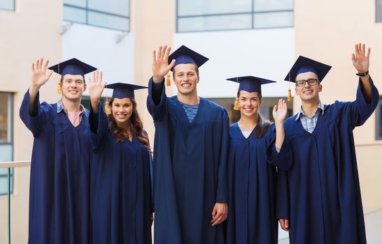 education, graduation and people concept - group of smiling students in mortarboards and gowns waving hands outdoors