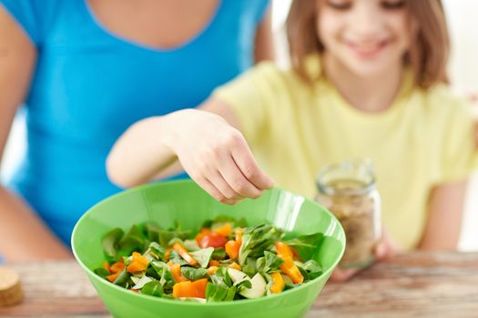 food, healthy eating, family and people concept - close up of happy girl and mother cooking salad for dinner and adding spices in kitchen