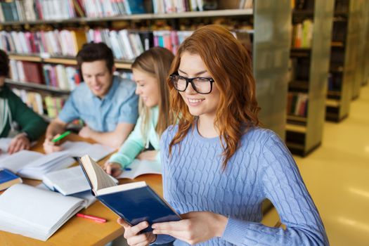 people, knowledge, education, literature and school concept - happy young woman in eyeglasses reading book and preparing to exams over group of students in library