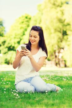 lifestyle, summer, vacation, technology and people concept - smiling young girl with smartphone sitting on grass in park
