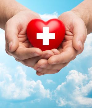 family health, charity and medicine concept - close up of male hands holding red heart with white cross over blue sky and clouds background