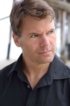 Closeup head shot of handsome Caucasian man in casual black shirt outdoors by pier at beach looking to camera right with intense expression of deep thought and focus
