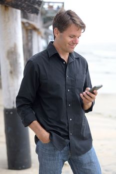 Hansome Caucasian man in casual clothes next to pier at beach on cloudy day smiles as he reads message on his smart phone