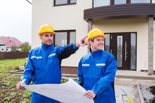 building, teamwork and people concept - two smiling builders in hardhats and overalls with blueprint pointing finger in front of house outdoors