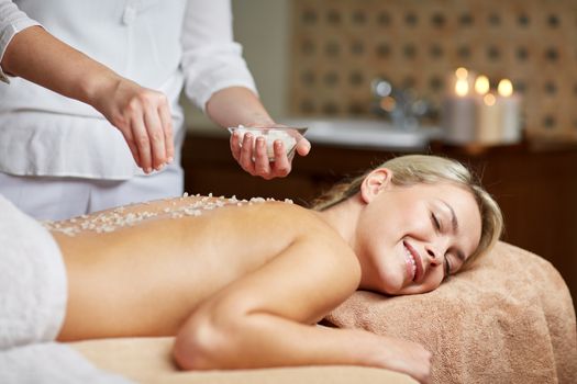 people, beauty, spa, massage and relaxation concept - close up of beautiful young woman lying with closed eyes and therapist holding salt bowl in spa