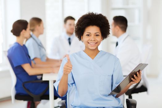 clinic, profession, people and medicine concept - happy female doctor or nurse with clipboard over group of medics meeting at hospital showing thumbs up gesture