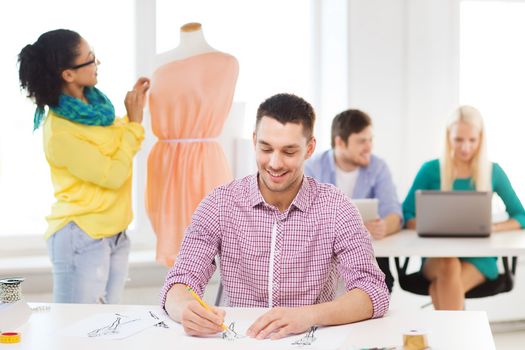 startup, education, fashion and office concept - smiling male drawing sketches and female adjusting dress on mannequin in office