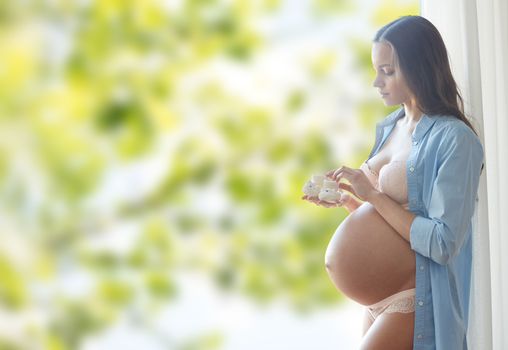 pregnancy, motherhood, people and expectation concept - happy pregnant woman with big bare tummy holding little baby booties at home over green natural background