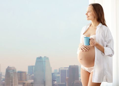 pregnancy, drinks, rest, people and expectation concept - happy pregnant woman in shirt and underwear with cup drinking tea over city background