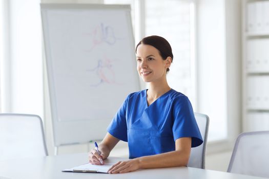 medicine, people and healthcare concept - happy female doctor or nurse writing medical report to clipboard at hospital