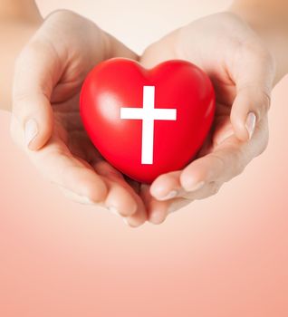 religion, christianity and charity concept - close up of female hands holding red heart with christian cross symbol over beige background