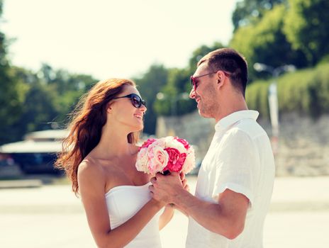 love, wedding, summer, dating and people concept - smiling couple wearing sunglasses with bunch of flowers looking at each other in city