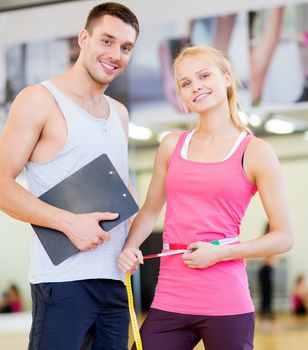 fitness, sport, training, diet, gym and lifestyle concept - two smiling people with clipboard and measure tape in the gym