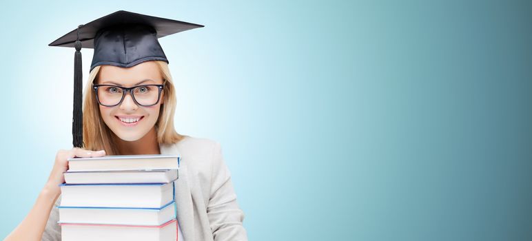 education, happiness, graduation and people concept - picture of happy student in mortar board cap with stack of books over blue background