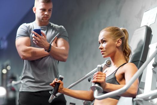 sport, fitness, bodybuilding, teamwork and people concept - young woman and personal trainer flexing muscles on gym machine