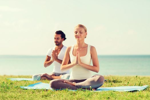 fitness, sport, friendship and lifestyle concept - smiling couple making yoga exercises sitting on mats outdoors