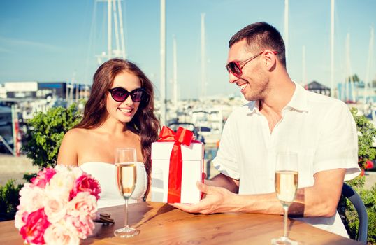 love, dating, people and holidays concept - smiling couple wearing sunglasses sitting with gift box, flowers and champagne at cafe