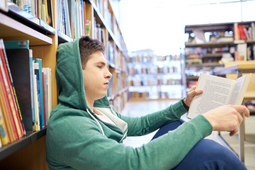 people, knowledge, education, literature and school concept - student boy or young man sitting on floor reading book in library