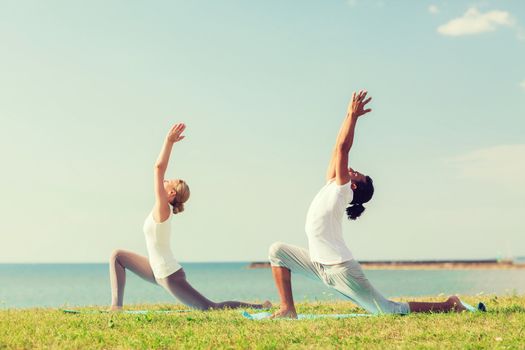 fitness, sport, friendship and lifestyle concept - smiling couple making yoga exercises on mats outdoors