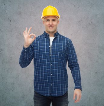 repair, construction, building, people and maintenance concept - smiling male builder or manual worker in helmet showing ok sign over gray concrete wall background