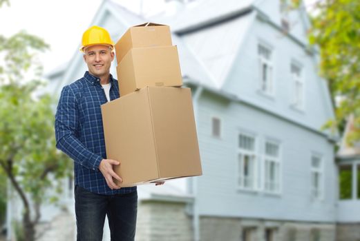 repair, building, construction, loading and delivery concept - smiling man or loader in helmet with cardboard boxes over living house background