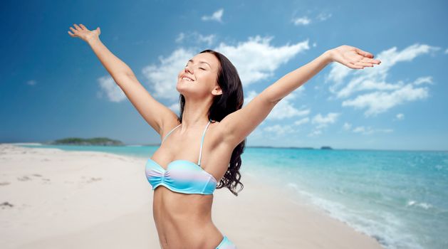 people, tourism, travel and summer concept - happy young woman in bikini swimsuit with raised hands over beach background