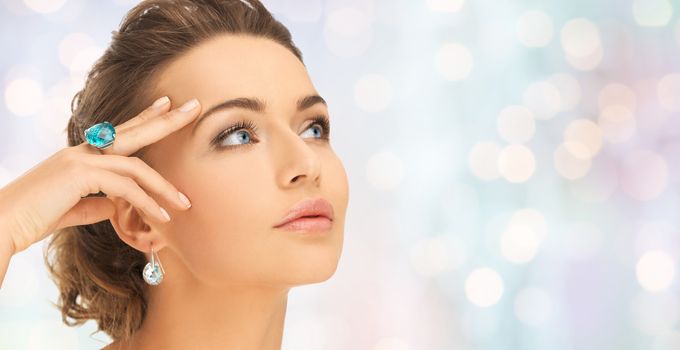 beauty, jewelry, people and accessories concept - close up of woman face with cocktail ring on hand and earrings over blue holidays lights background