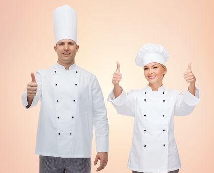 cooking, profession, gesture, teamwork and people concept - happy chefs or cooks couple showing thumbs up over beige background