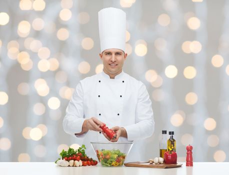 profession, vegetarian, food and people concept - happy male chef cooking and seasoning vegetable salad over christmas holidays lights background