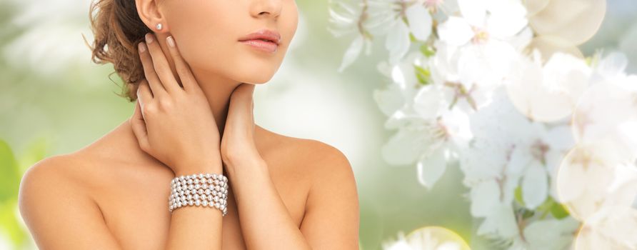 beauty, people and jewelry concept - close up of beautiful woman with pearl earrings and bracelet over summer garden and cherry blossom background