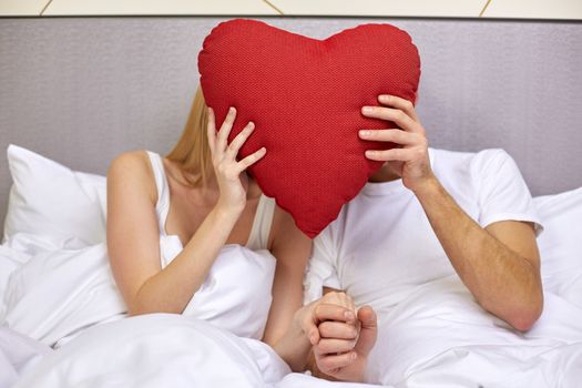 travel, love, valentines day, holidays and happiness concept - happy couple in bed hiding faces behind red heart shape pillow at hotel or home